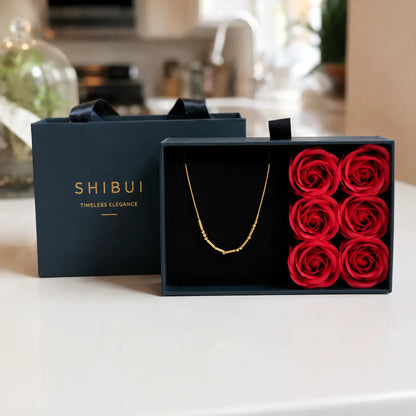 A gold necklace presented in an open black gift box next to a matching black box lid, both printed with the brand name SHIBUI. The gift box is styled with six red preserved roses on a white countertop in a blurred kitchen setting.
