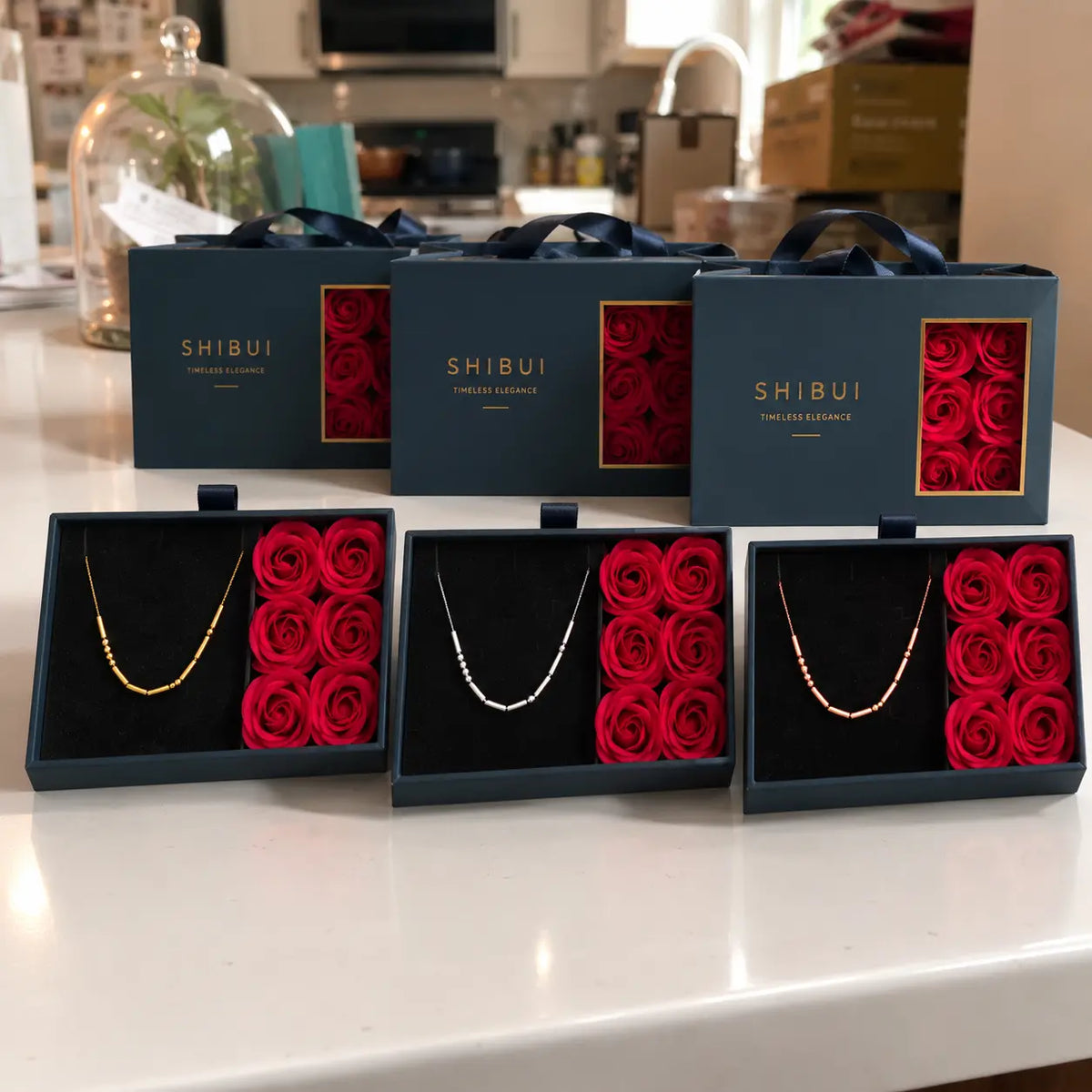 Three open SHIBUI jewelry gift boxes on a white countertop, each containing a delicate necklace beside a set of deep red rose-shaped cushions, with three closed matching boxes behind them.