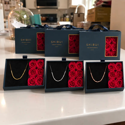 Three open SHIBUI jewelry gift boxes on a white countertop, each containing a delicate necklace beside a set of deep red rose-shaped cushions, with three closed matching boxes behind them.